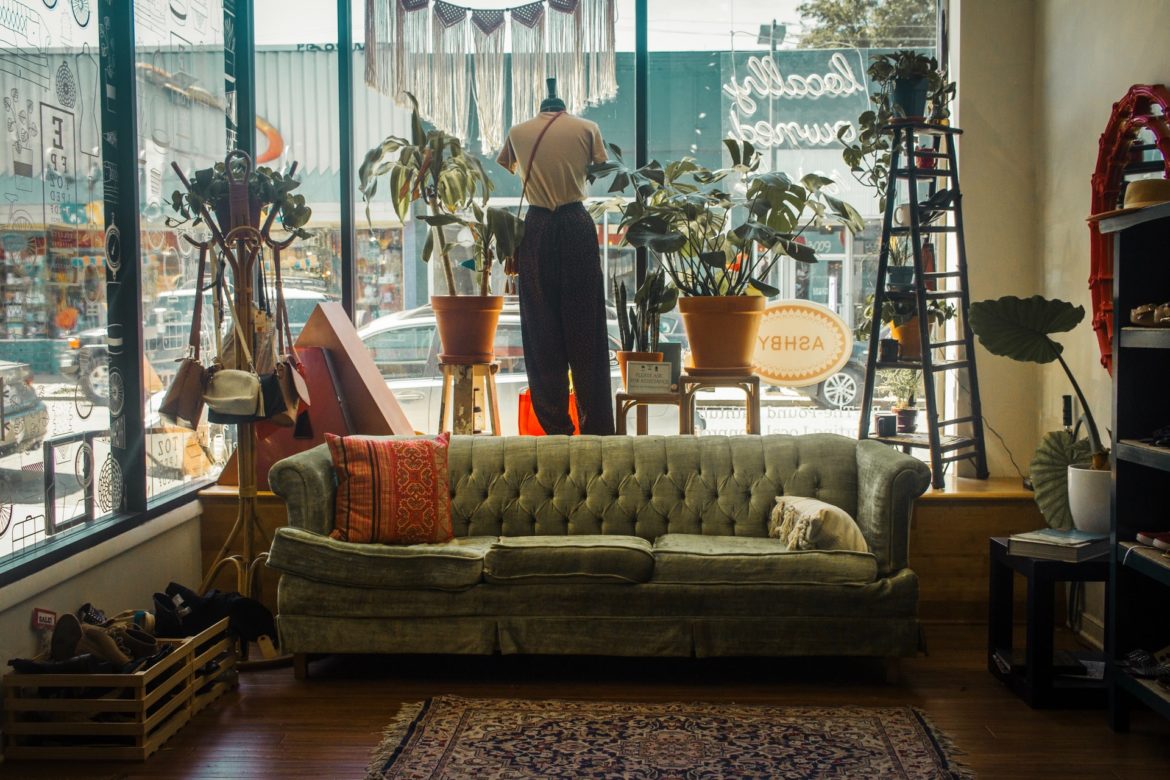 Grünes Sofa in einem Second Hand Shop vor dem Fenster, eingerahmt von alten Lampen und Mannequins
